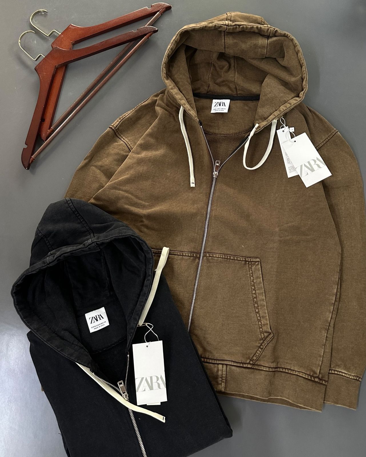 Olive brown and washed black zip hoodies laid flat next to wooden hangers